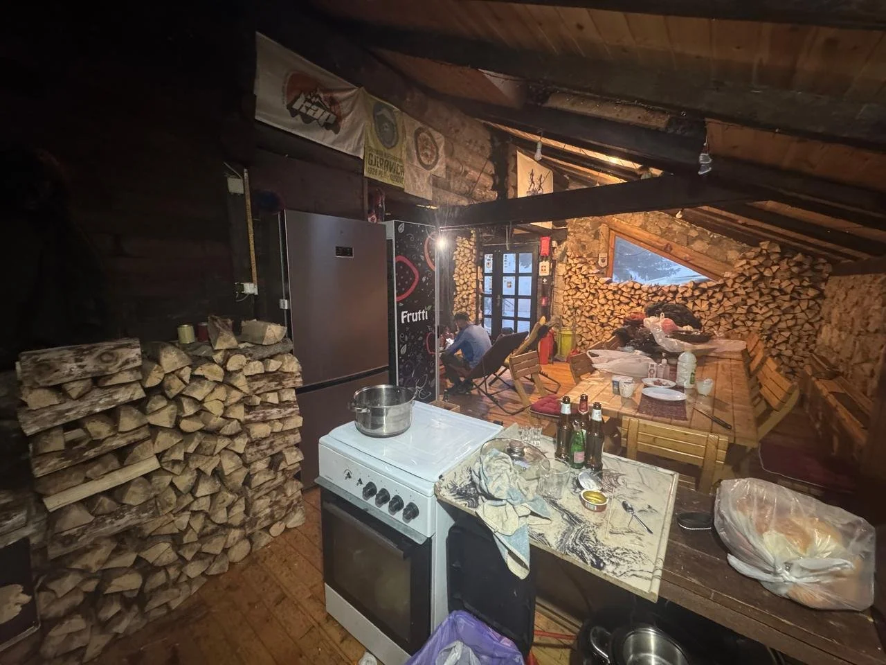Inside a rustic cabin kitchen with wood walls, firewood stacked on left and back walls, a small stove with a pot, a table with bottles, dishes, and food, a refrigerator, and a person sitting in a chair in the background near a window.