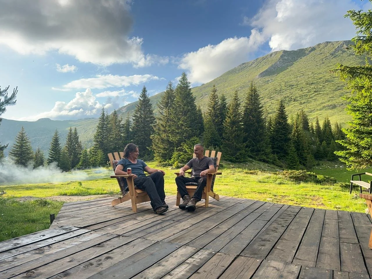 Two people sitting in wooden Adirondack chairs on a wooden deck outdoors, surrounded by green grass, pine trees, and mountains under a partly cloudy sky.