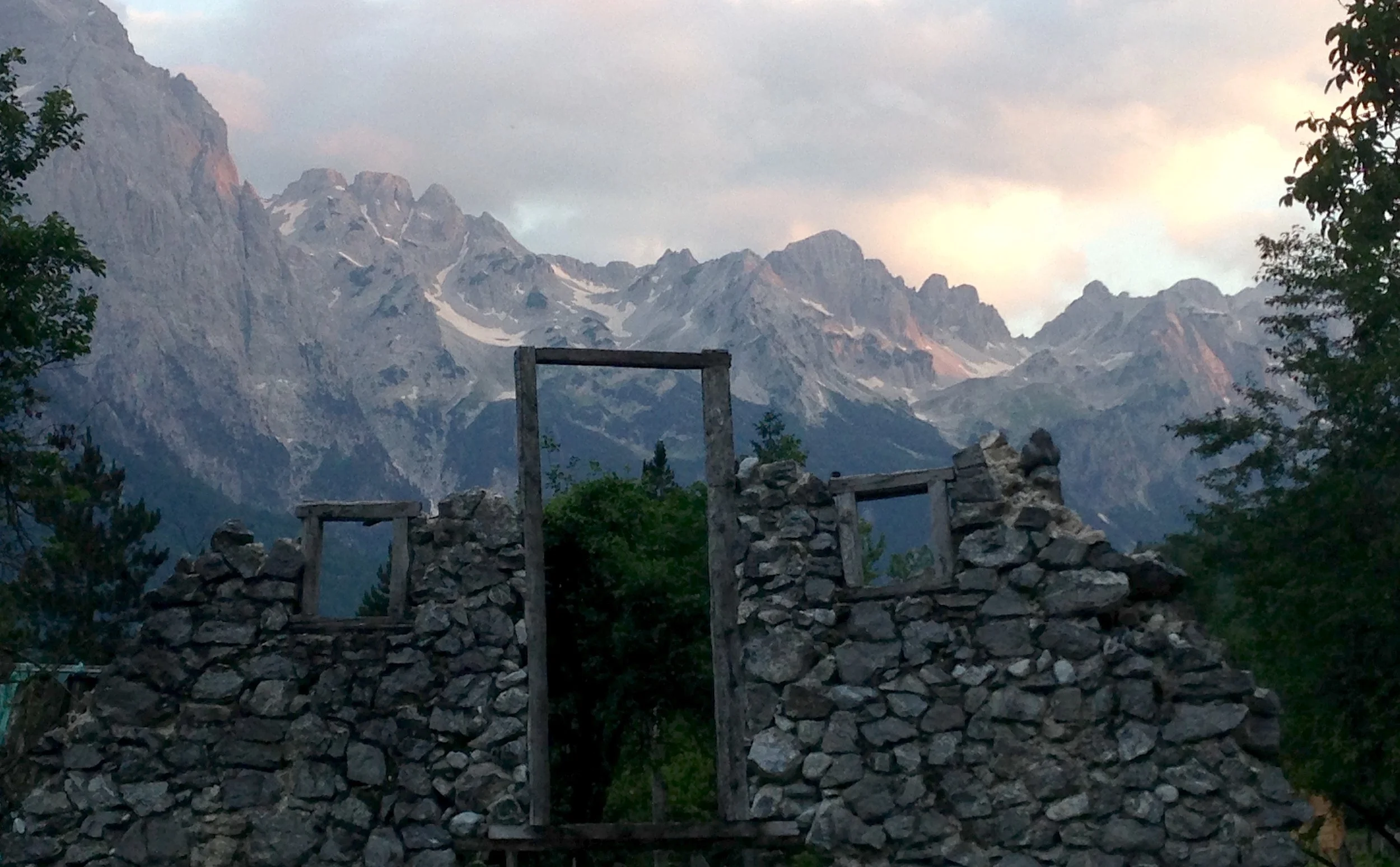 A mountain landscape with snow-capped peaks, trees, and a stone wall or ruins in the foreground.