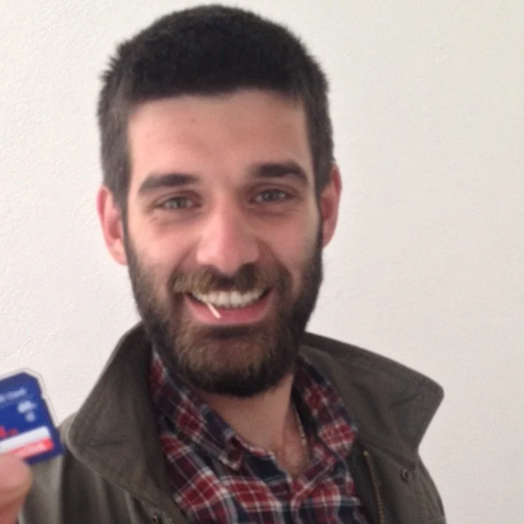 A young man with a beard and short dark hair smiling at the camera, wearing a plaid shirt and a dark jacket, against a plain light-colored wall.