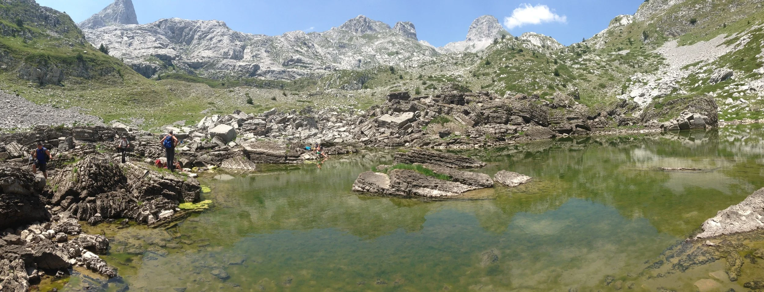 Group of hikers exploring a rocky lakeside in a mountainous area with green hills and rugged peaks under a clear blue sky.