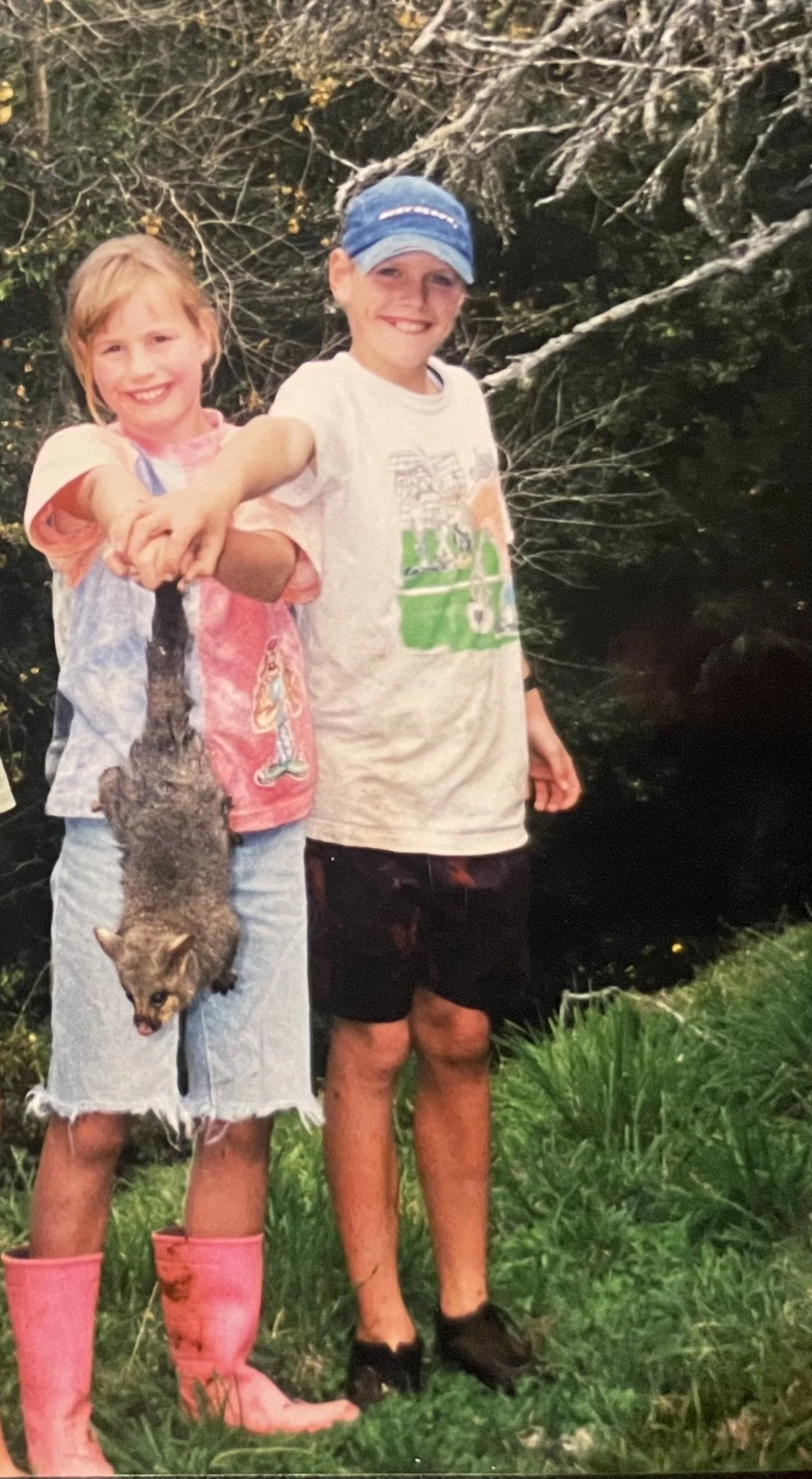 Marcy and Owen as children, standing in a paddock, next to a stream with greenery and trees, holding a possum together.
