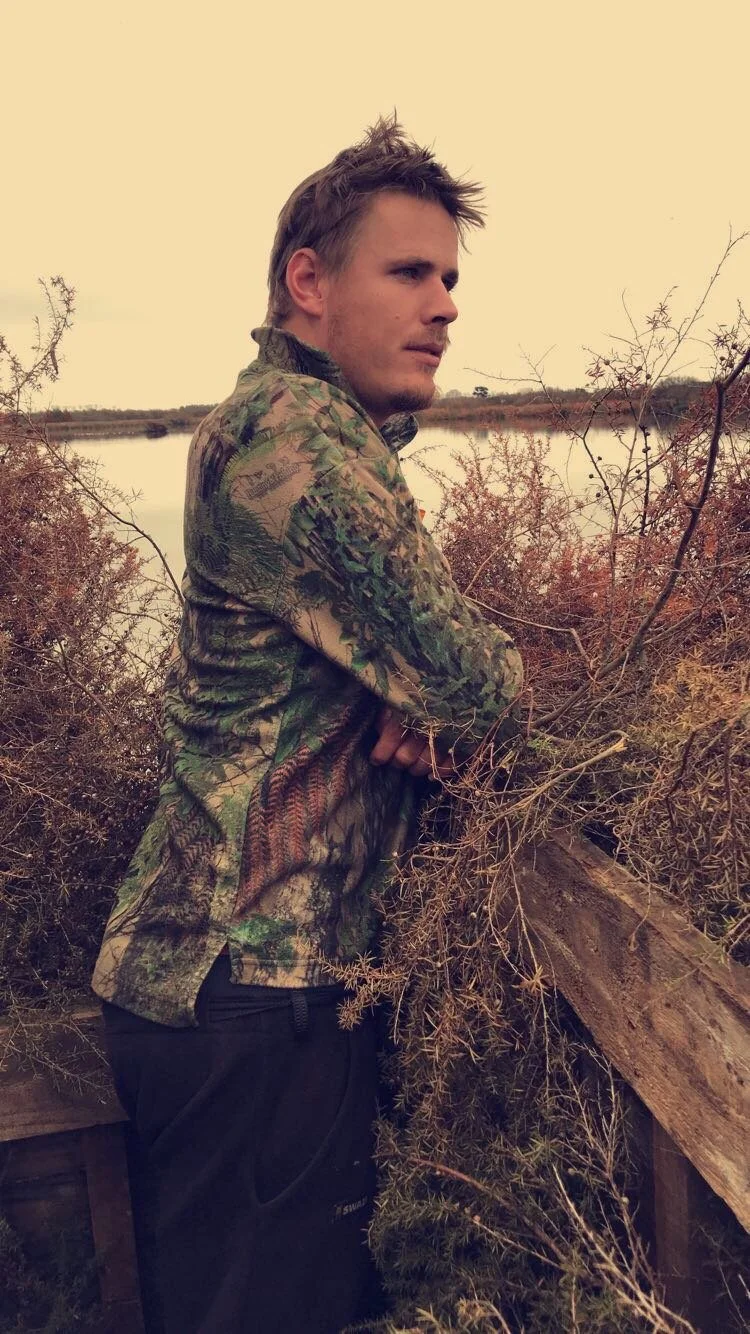 Owen Bakker standing in a Maimai near a body of water at a lake in Huntly, looking to the side with a contemplative expression.