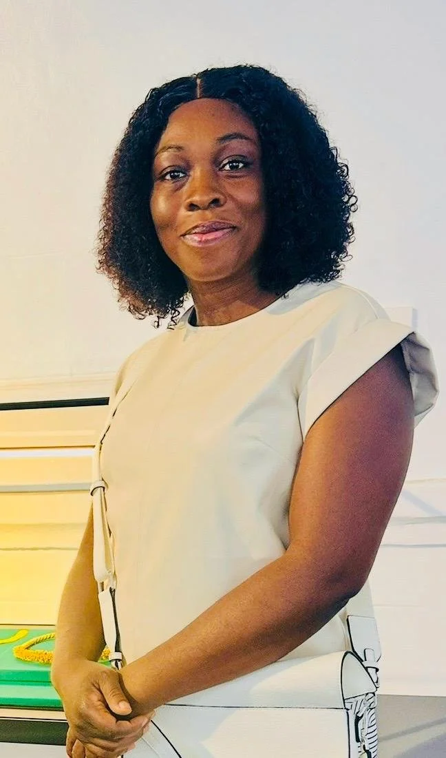 A woman with curly black hair wearing a light-colored dress, standing indoors with a slight smile.
