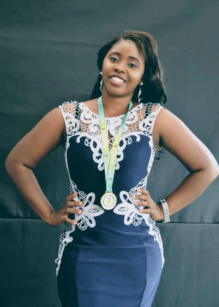 Woman in navy blue dress with white lace details, smiling, wearing a medal around her neck and a bracelet, standing with hands on hips against a dark background.