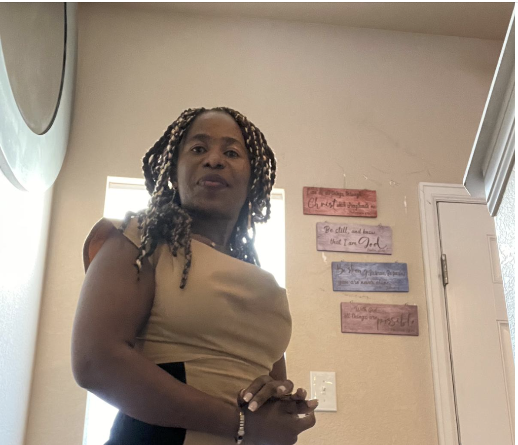 A woman with braided hair standing indoors with sunshine behind her, next to a wall with four colorful decorative signs.