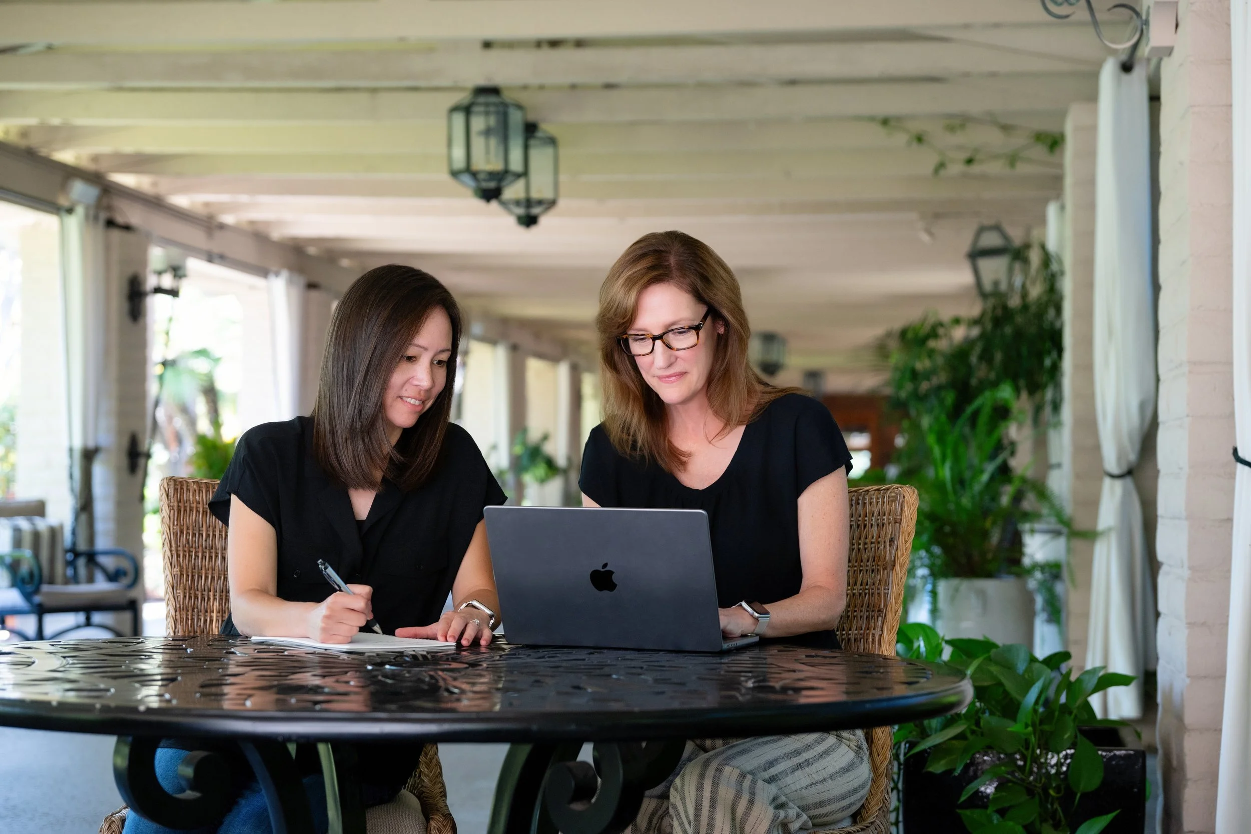 Two women sit together at an outdoor patio table, looking at a laptop and writing notes. The scene is bright and inviting, with lantern-style lights hanging from a wooden ceiling and greenery in the background.