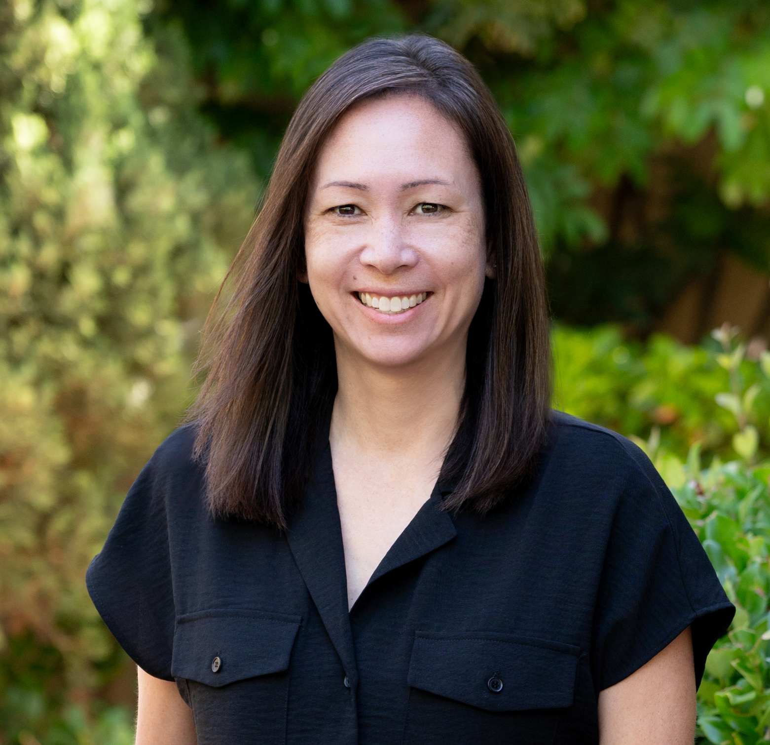 A woman with long brown hair smiling outdoors in front of a green, leafy background, wearing a black shirt.