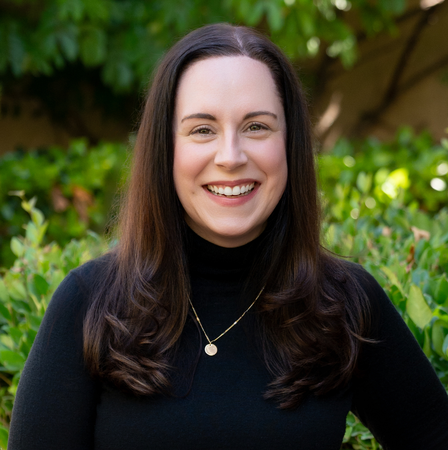 A woman with long dark hair smiling outdoors, wearing a black turtleneck and gold jewelry, with green leafy background.