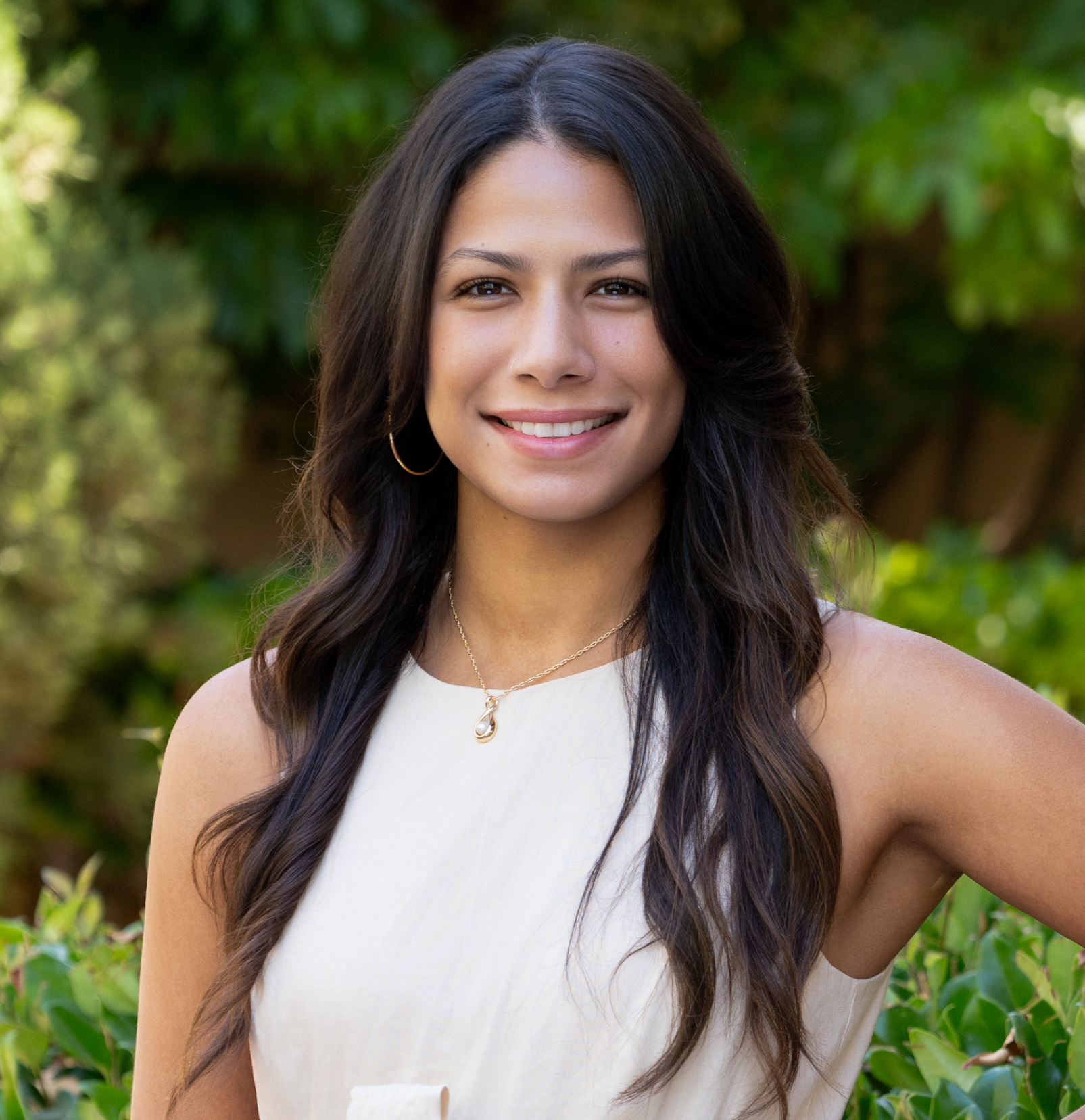 A young woman with long dark wavy hair, wearing a white sleeveless top, gold hoop earrings, and a gold necklace with a pearl pendant, standing outdoors with greenery in the background and smiling.