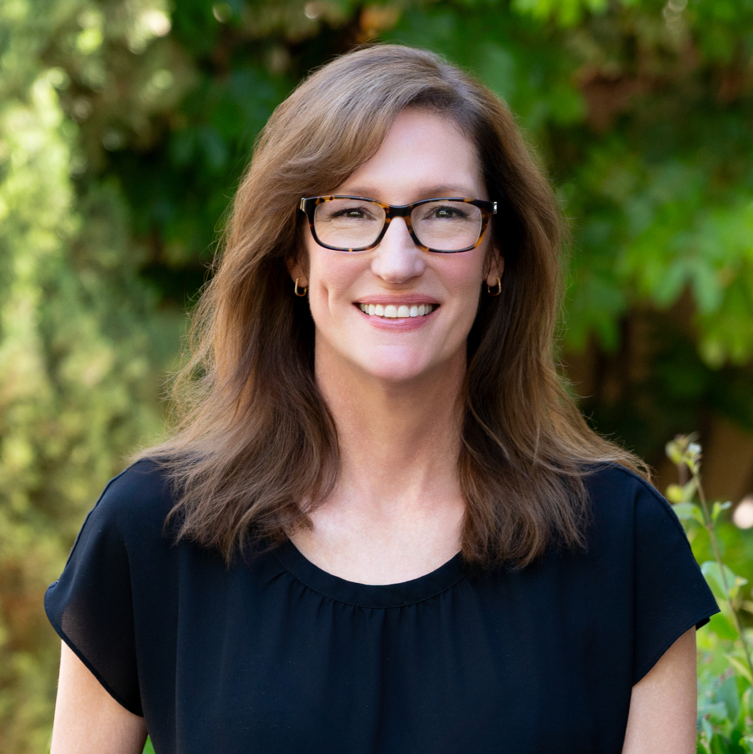 A woman with shoulder-length brown hair, wearing glasses and gold hoop earrings, smiling outdoors with a background of green foliage.