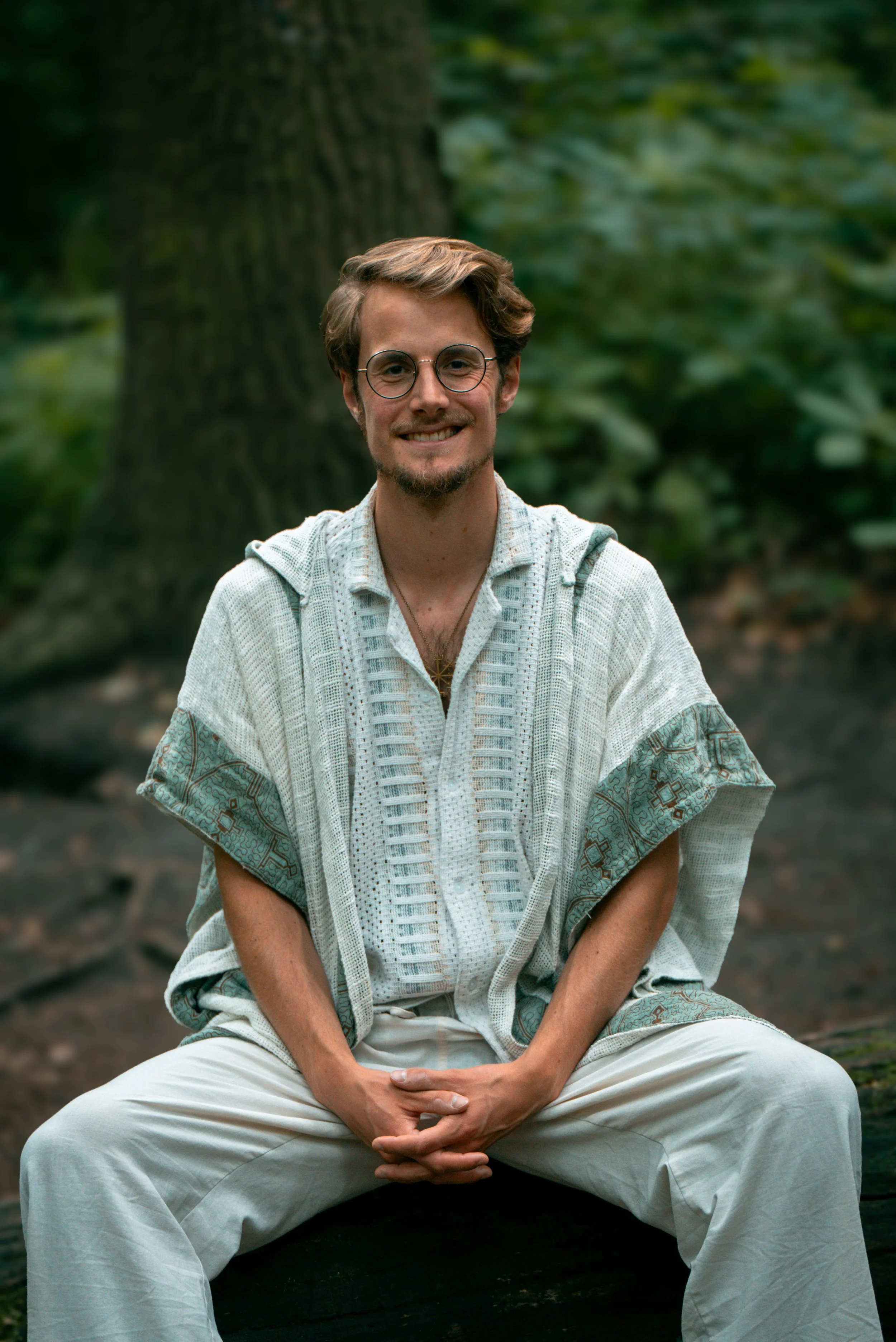 Ein Mann mit Brille und lockigem Haar sitzt vor einem Baum in einem grünen Wald und lächelt.