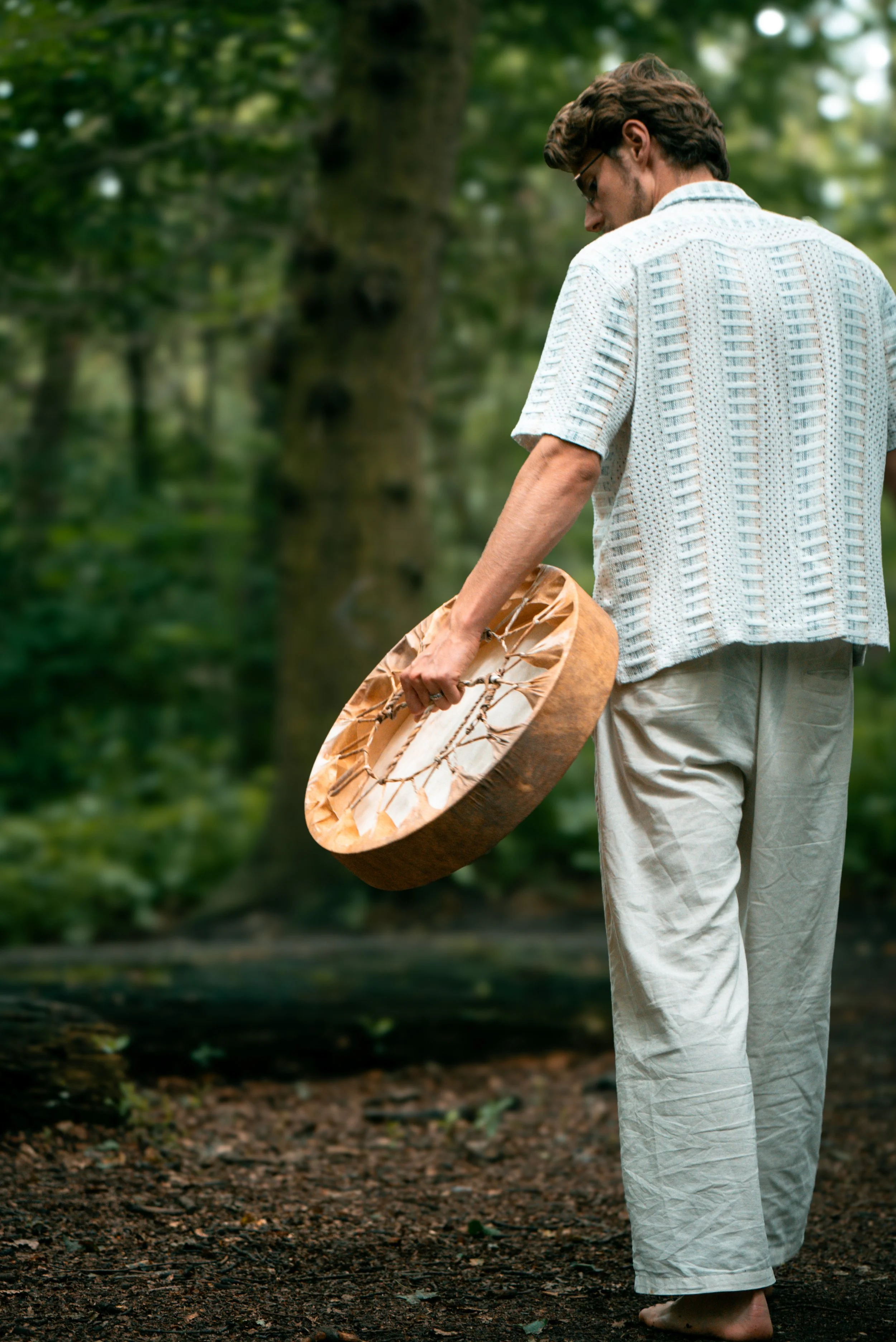 Ein Mann in weißen lockeren Kleidern steht barfuß im Wald und hält ein handgefertigtes, rundes Trommelinstrument aus Holz in der Hand.
