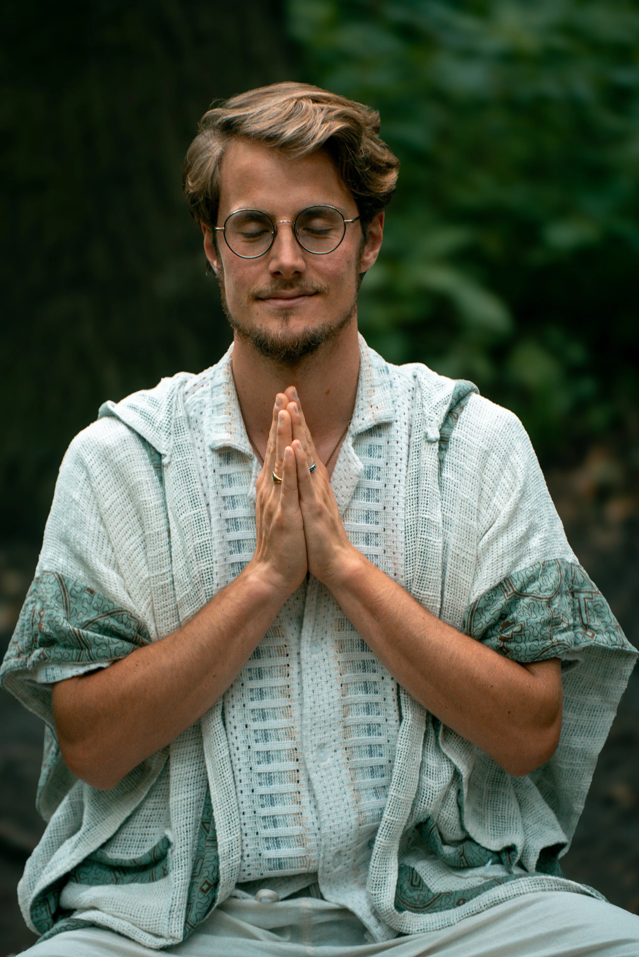 Ein junger Mann mit Brille und Bart sitzt im Freien, die Hände in Gebetshaltung, mit geschlossenen Augen, in einer meditativen Pose.