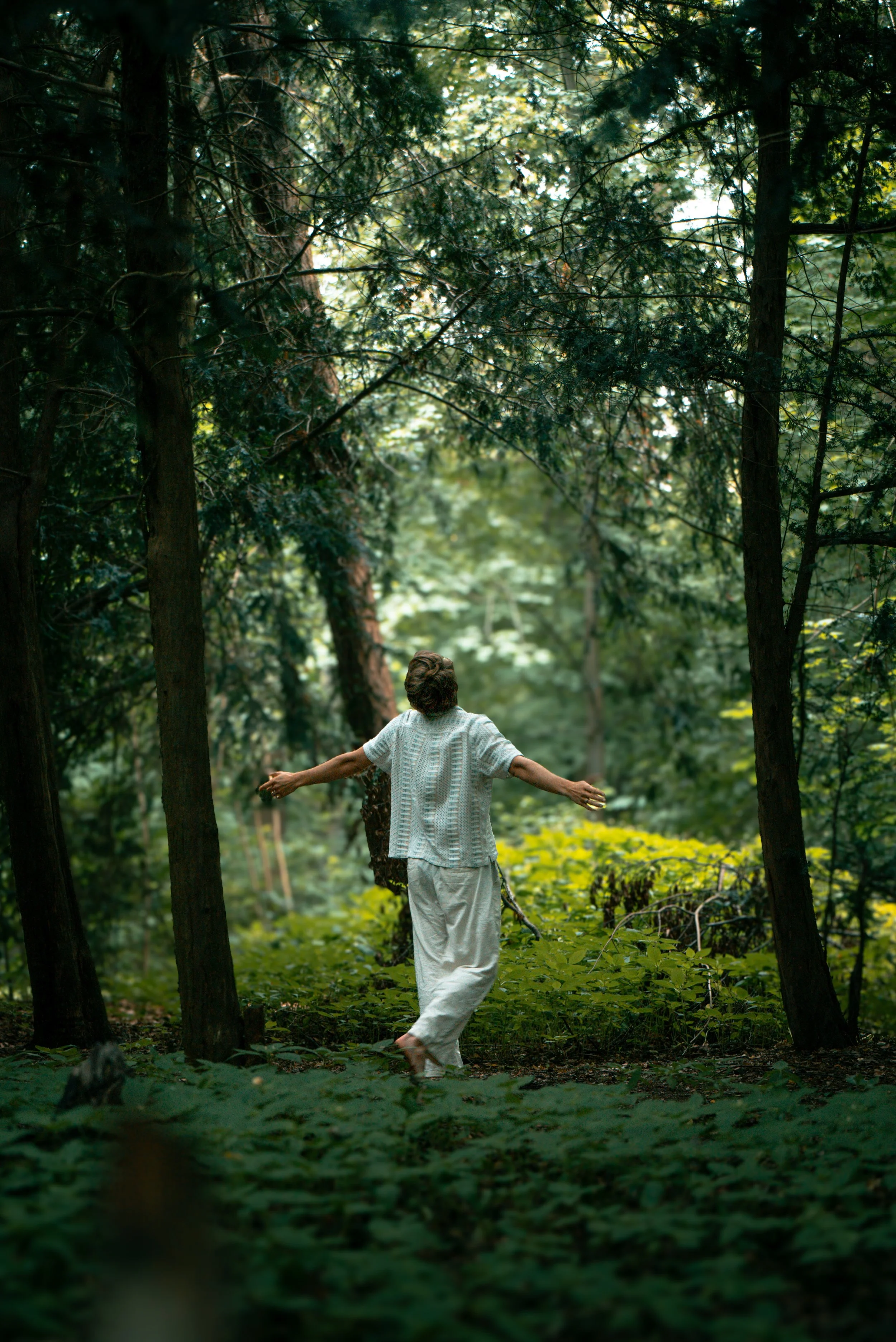 Eine Person in einem weißen Outfit tanzt barfuß in einem dichten, grünen Wald.