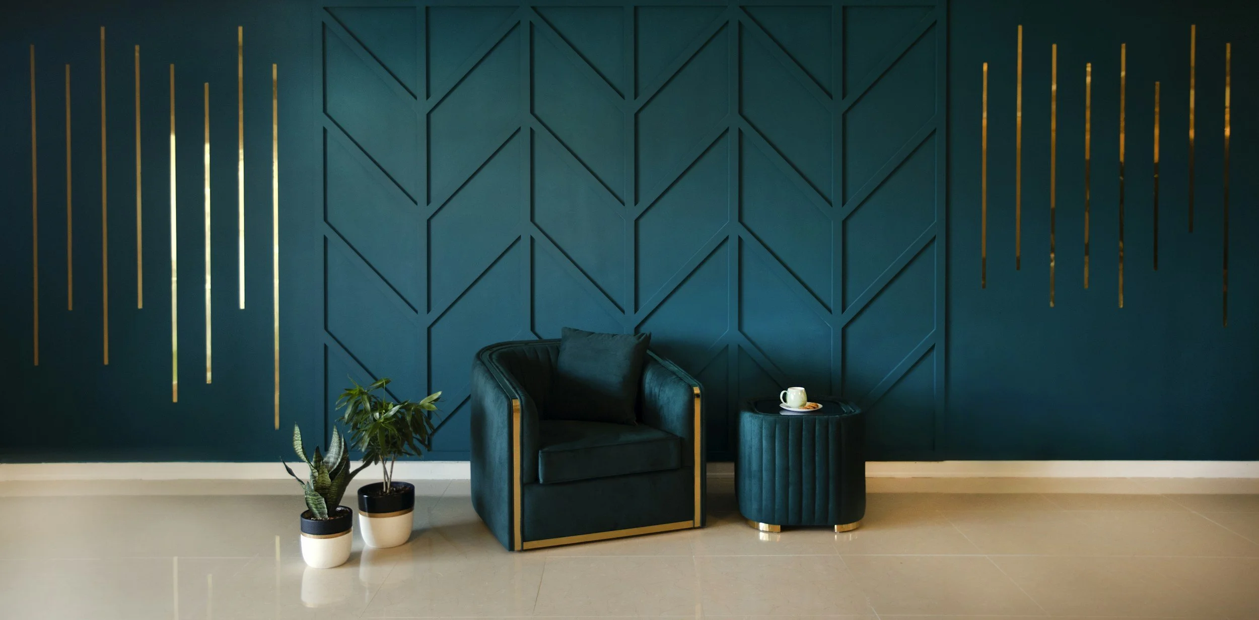 A modern interior with a dark teal accent wall featuring geometric paneling and gold vertical decorative strips, a dark velvet armchair with a pillow, a matching round ottoman, a small side table with a white cup and saucer, and potted plants on the floor.