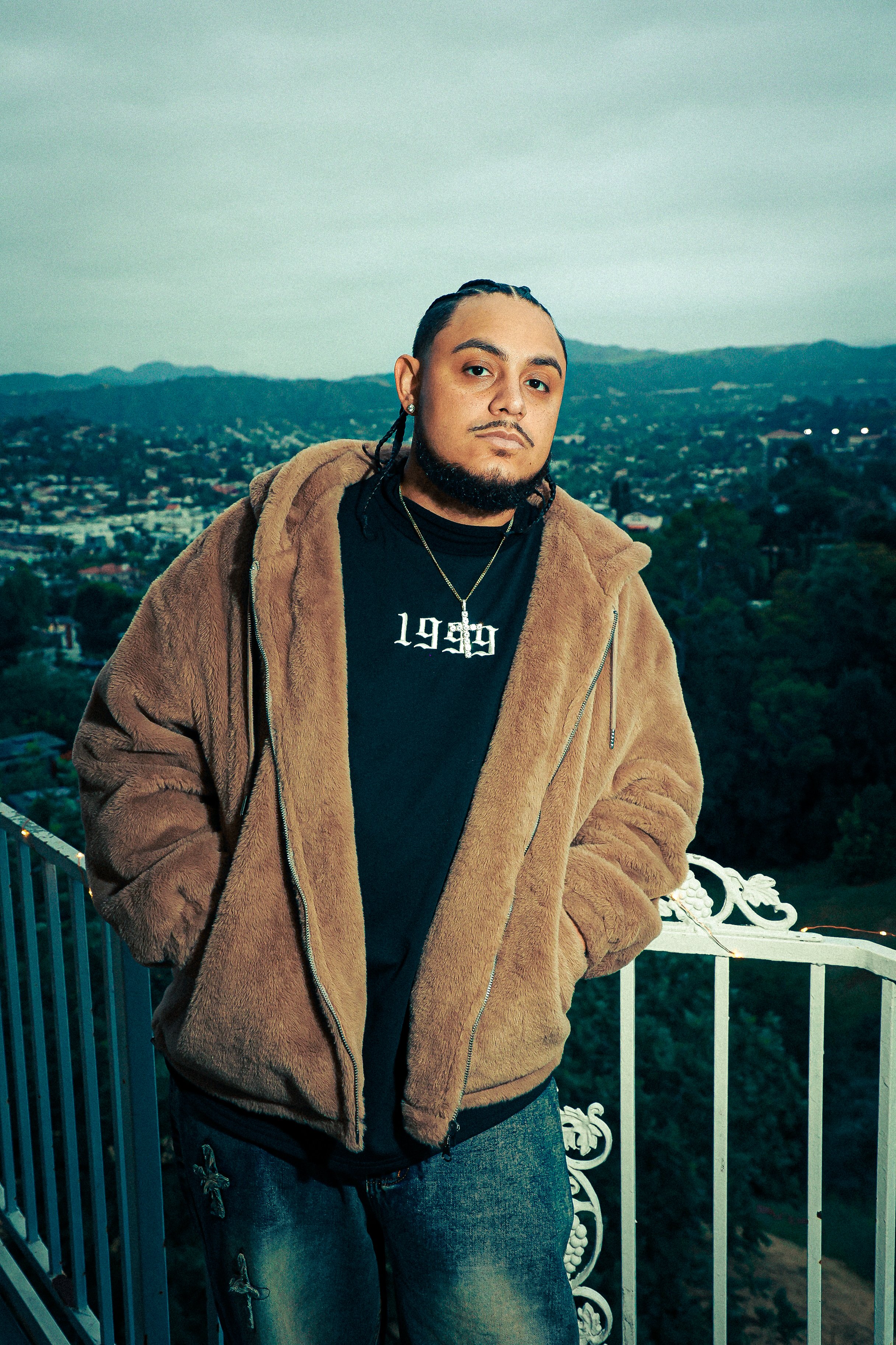 A young man with braided hair and a beard, wearing a brown fuzzy jacket, a black T-shirt with white text, jeans with chain accessories, and jewelry, stands on a balcony with a scenic city and mountain view in the background during twilight.
