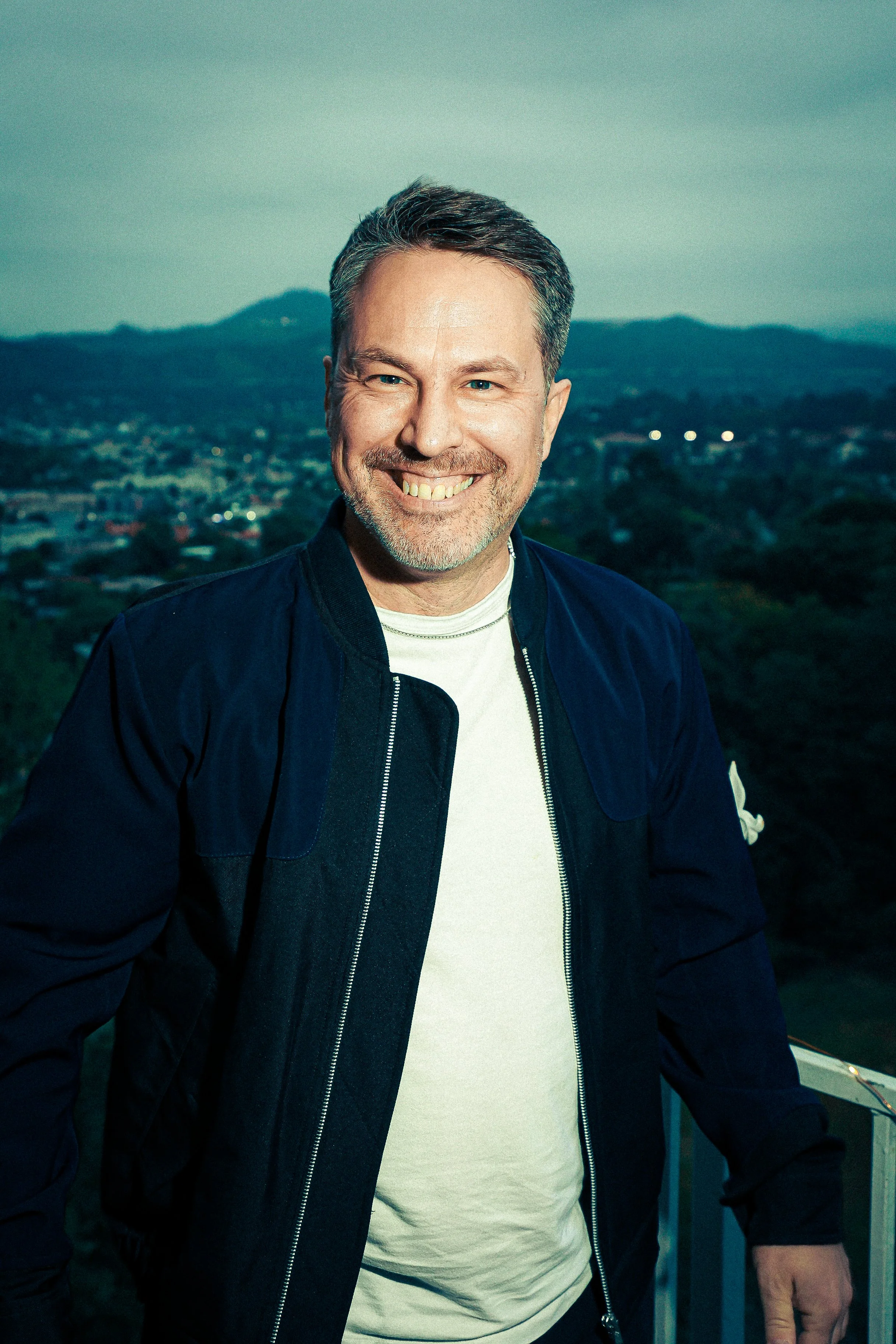A smiling man with short, dark hair and a beard, wearing a dark jacket over a white shirt, standing outdoors during dusk with mountains and trees in the background.