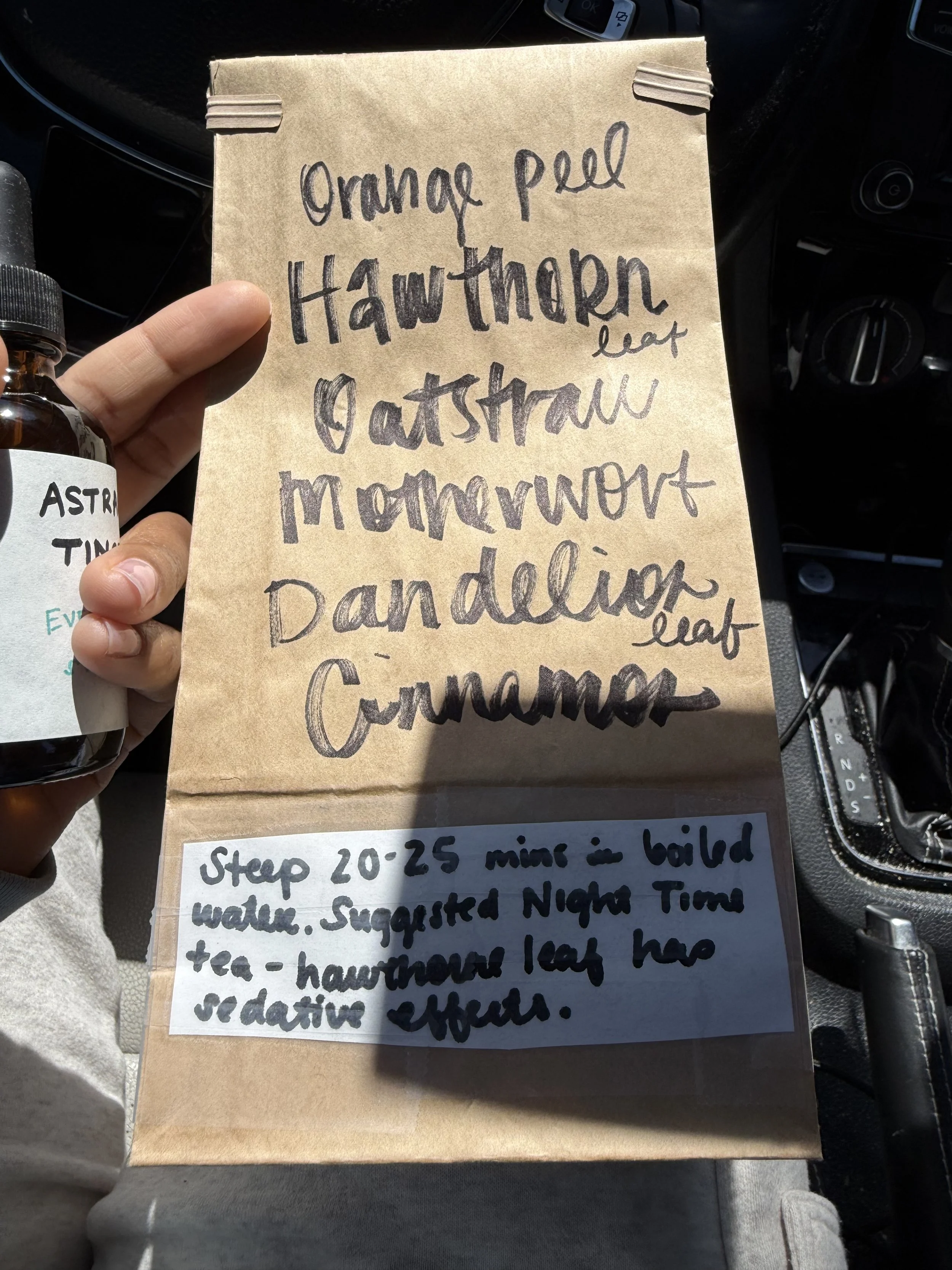 A hand holding a paper bag of herbal tea with handwritten notes listing items like orange peel, hawthorn leaf, oatstraw, cinnamon, oatstraw.
