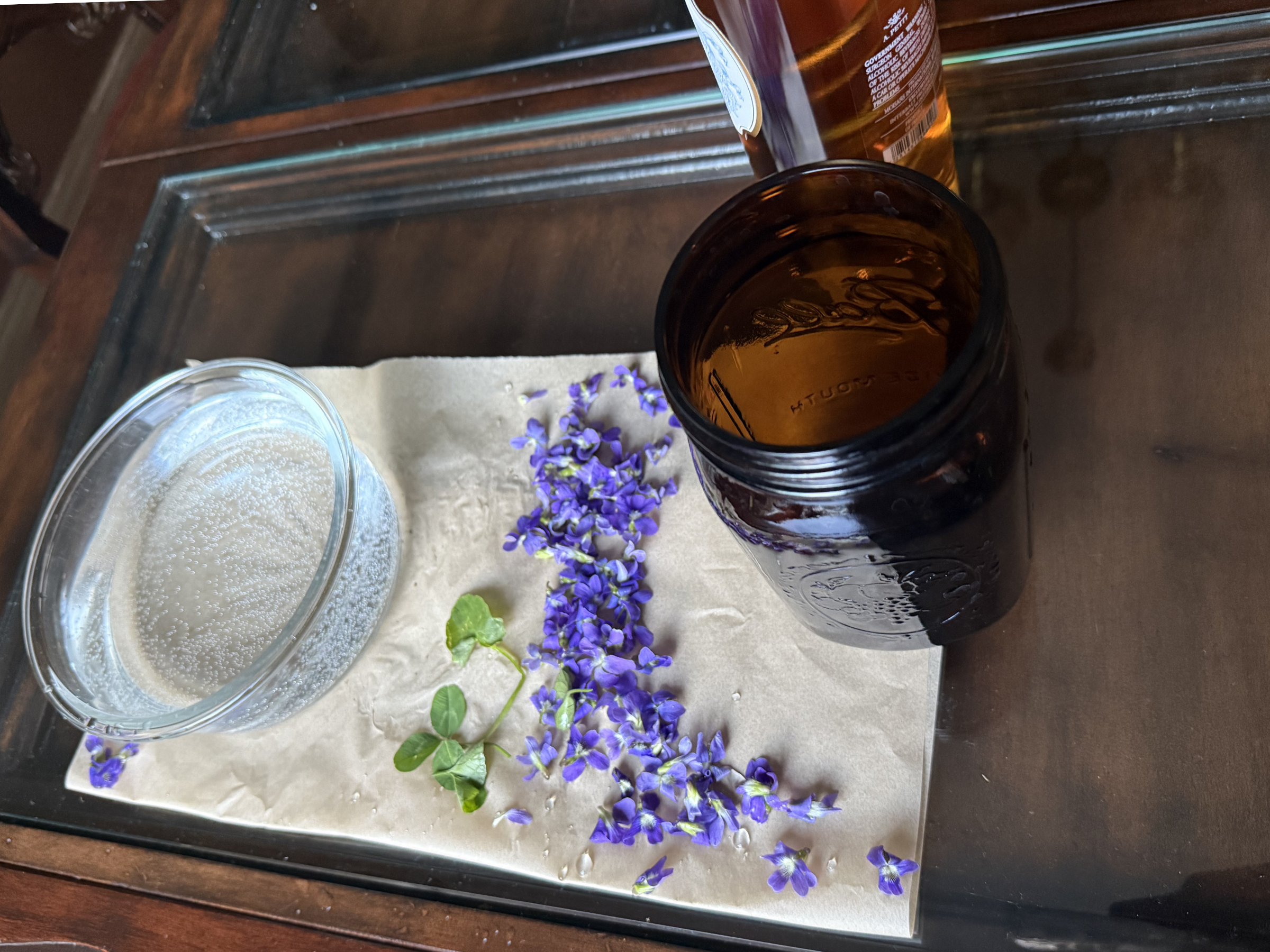 A glass of clear sparkling water, a jar with amber liquid, purple flowers, and green leaves on a table for making flower essences.