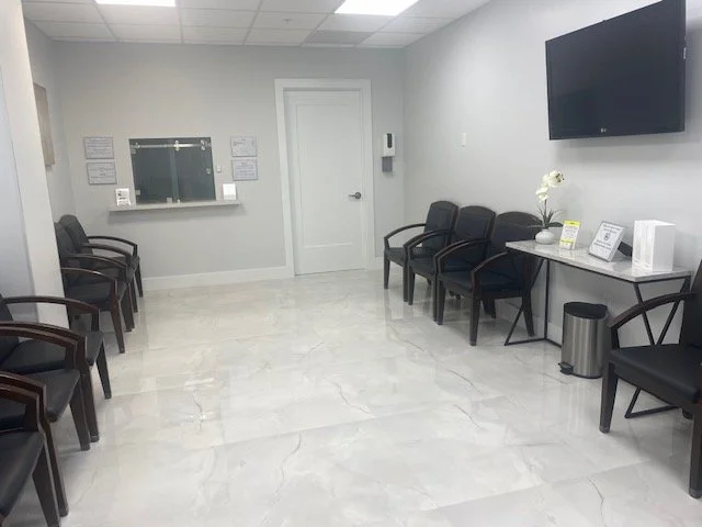 Empty waiting room with black chairs, a white table with flowers, a large TV, and a window on the wall at a healthcare facility.