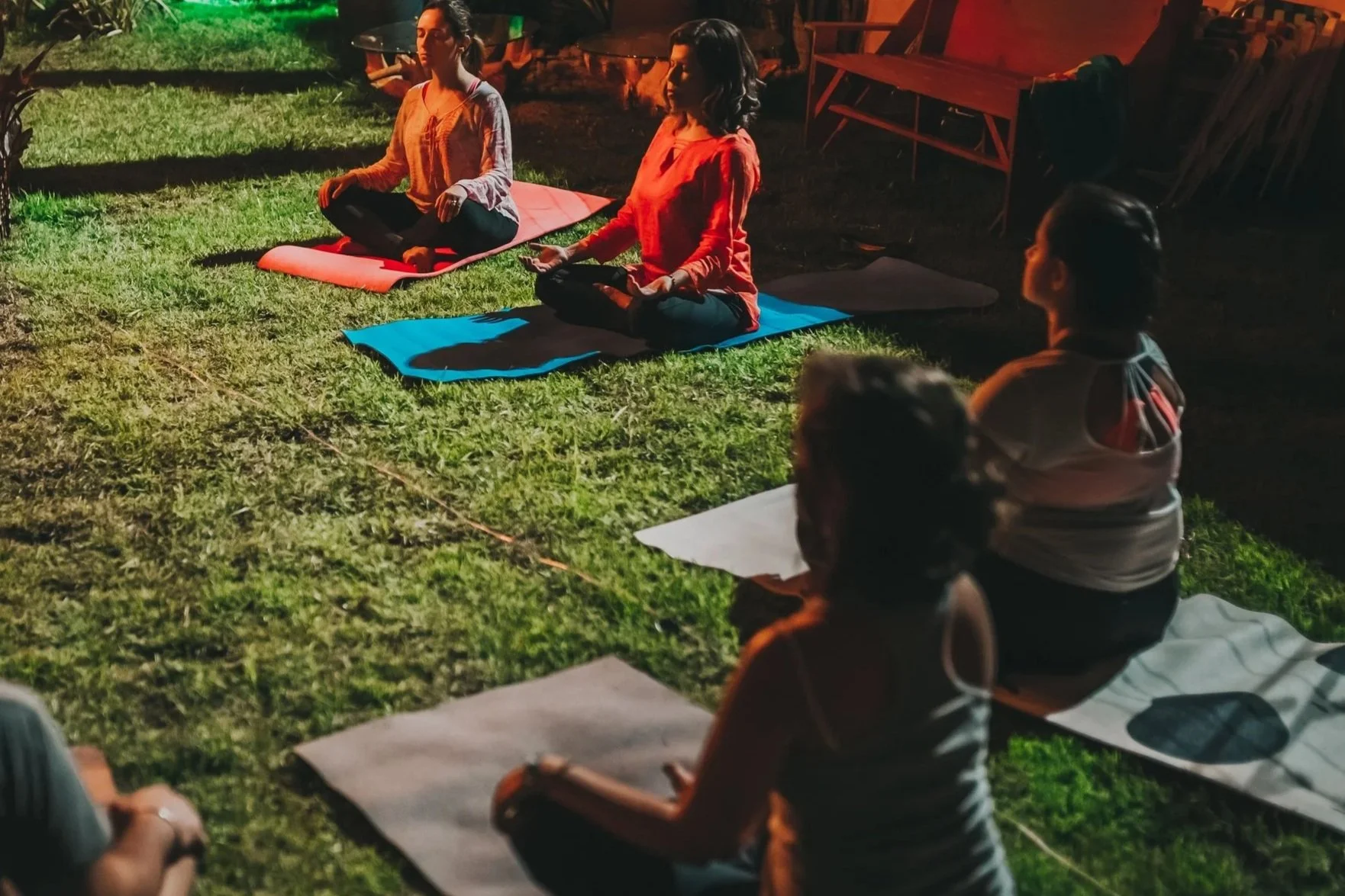 Group of women practicing yoga outdoors at night, sitting cross-legged on yoga mats with their eyes closed and hands resting on their knees.