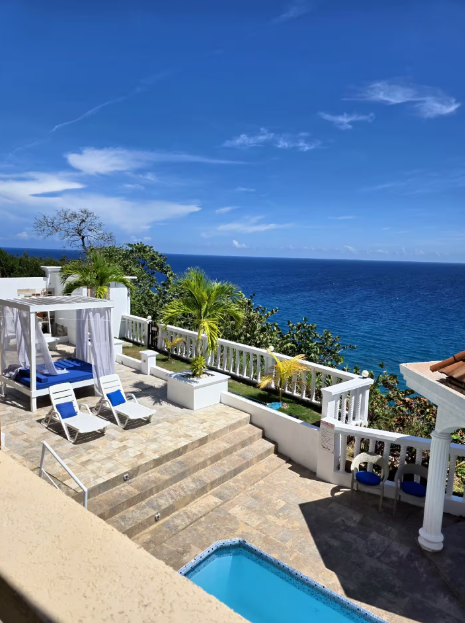 Outdoor terrace overlooking the ocean with lounge chairs, a small pool, and lush greenery under a blue sky.