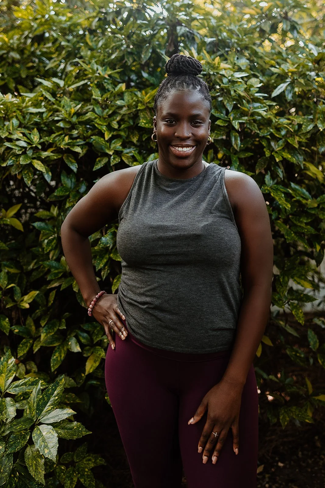 A woman with dark hair in a bun, wearing a gray sleeveless top and maroon pants, standing outdoors in front of green leafy bushes, smiling with one hand on her hip.