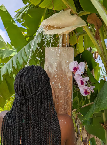 Person with braided hair stands in front of a wooden shower with water pouring from a cactus-shaped spout, surrounded by lush green banana leaves and purple orchids.