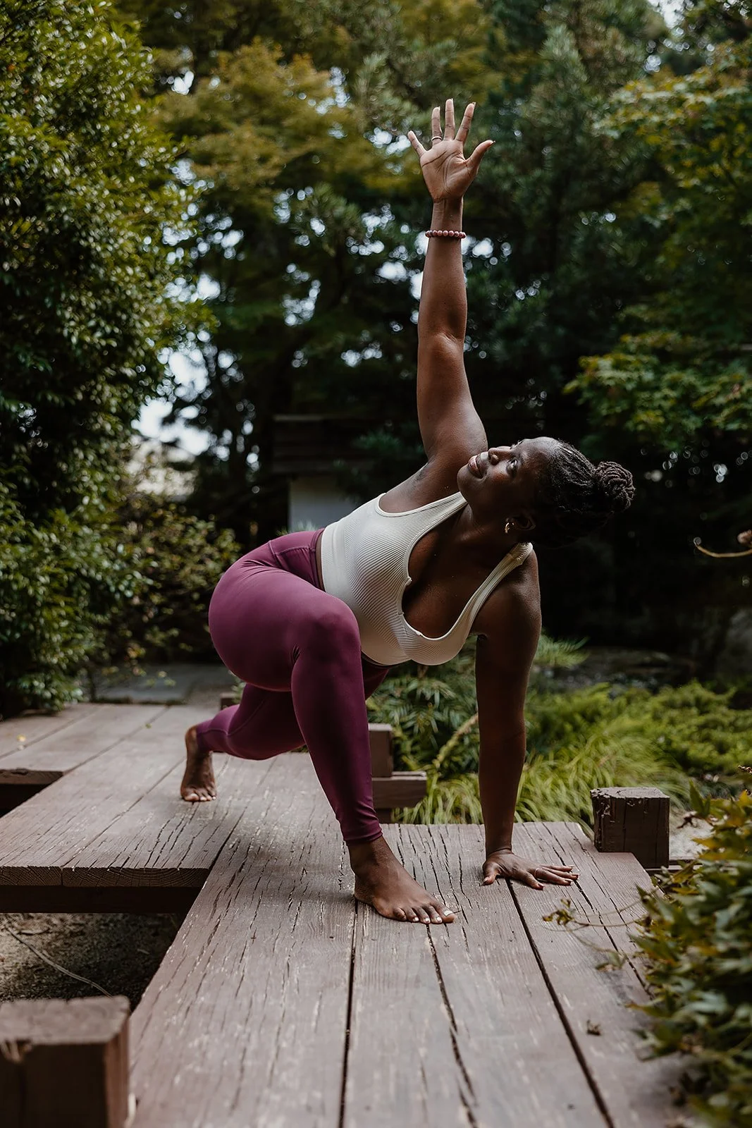 A woman practicing yoga outdoors on a wooden deck surrounded by greenery, in a pose with one hand on the deck and the other reaching upwards.