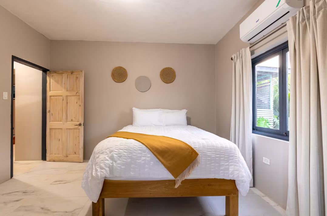 A minimalist bedroom with a wooden bed frame, white bedding, a yellow throw blanket, and decorative woven wall hangings. The room has beige walls, a window with white curtains, and an air conditioning unit.