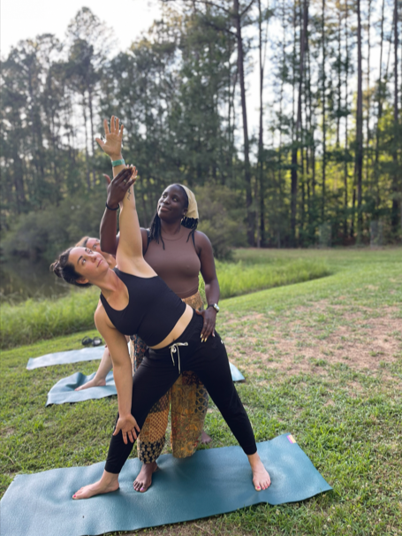Two women practicing yoga outdoors in a grassy field with trees in the background. One woman in black is in a side bend pose on a yoga mat, while the other woman stands behind her, assisting and observing.