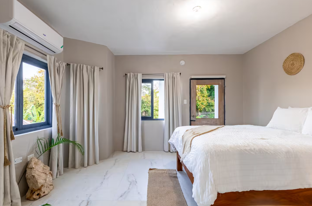 Bright bedroom with white bedding, beige walls, and large windows with cream curtains, wooden door, and minimal decor.