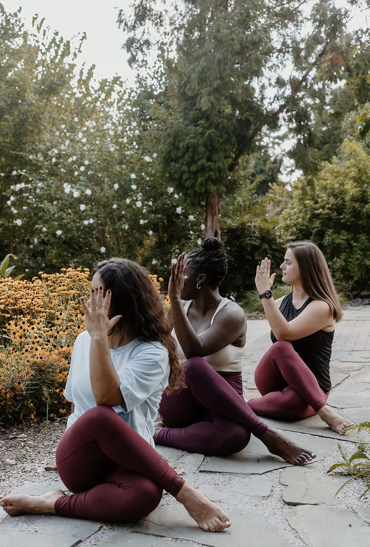 Three women practicing yoga outdoors, sitting cross-legged on a stone path, with trees and yellow flowers in the background.