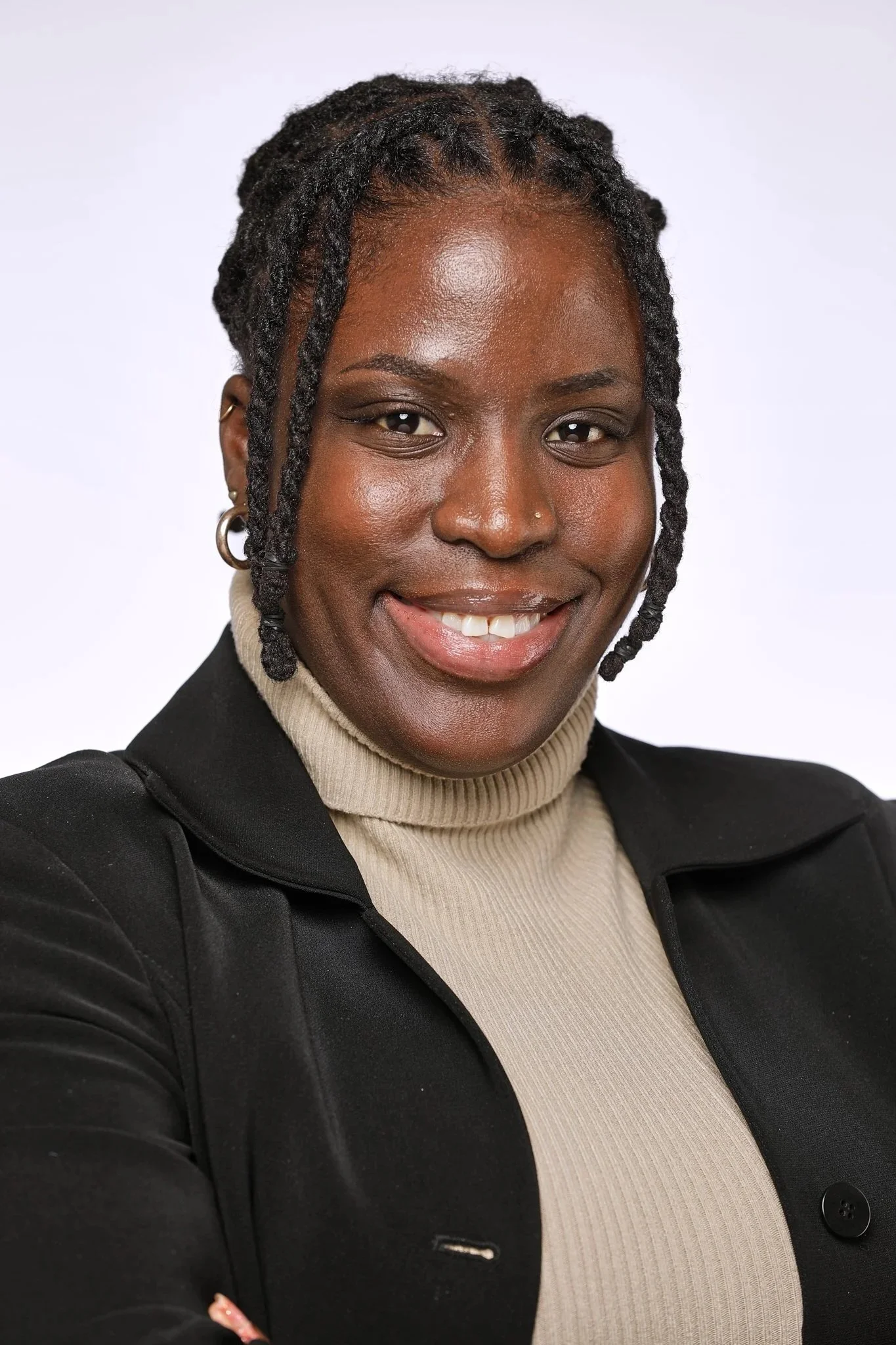 Close-up photo of an African American woman with dreadlocks, smiling, wearing a beige turtleneck and a black blazer, standing against a white background.