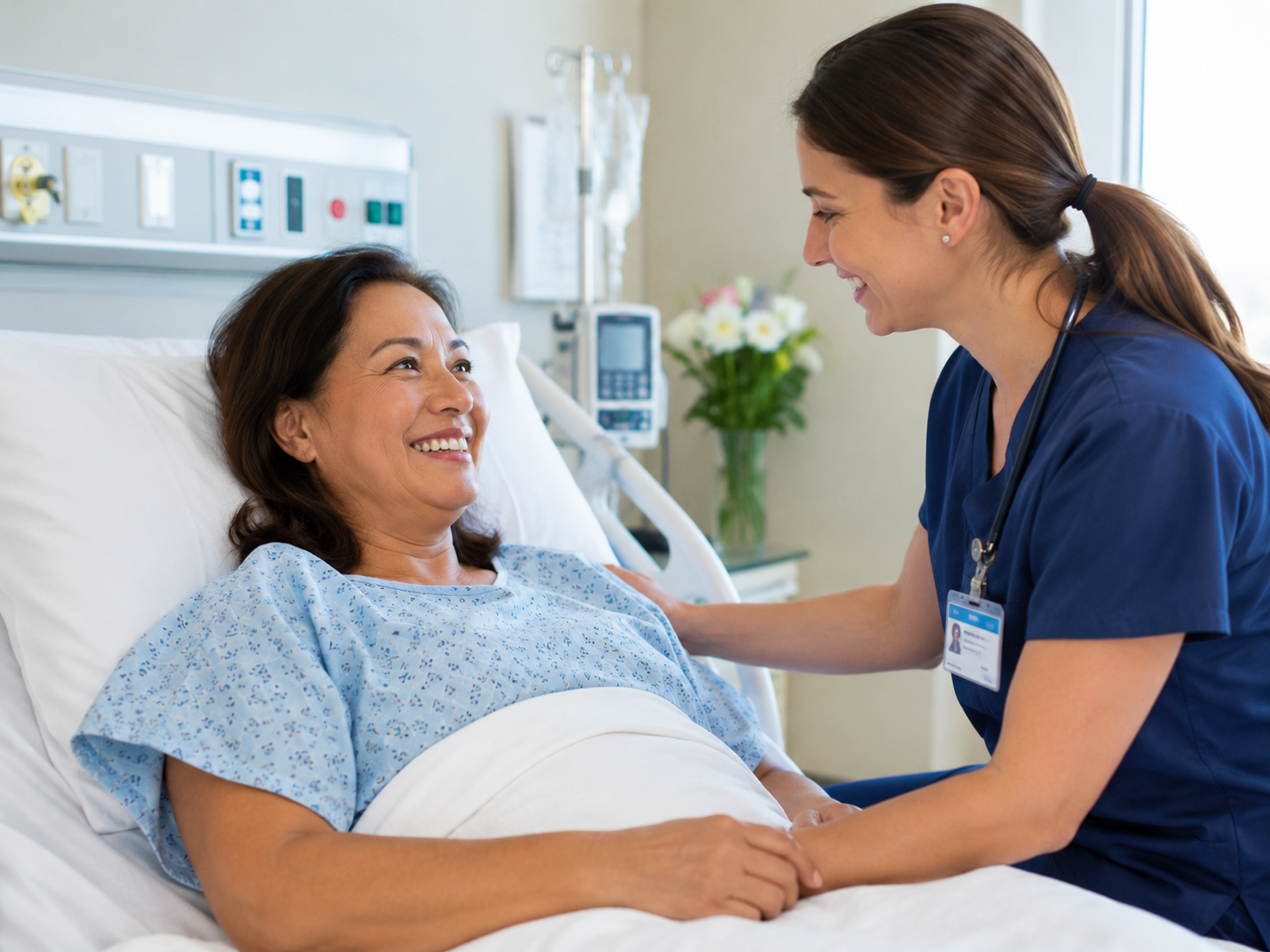 A nurse in blue scrubs with a name badge smiling and talking to a woman lying in a hospital bed, holding her hand. The patient is smiling back, wearing a hospital gown, with medical equipment and flowers in the background.
