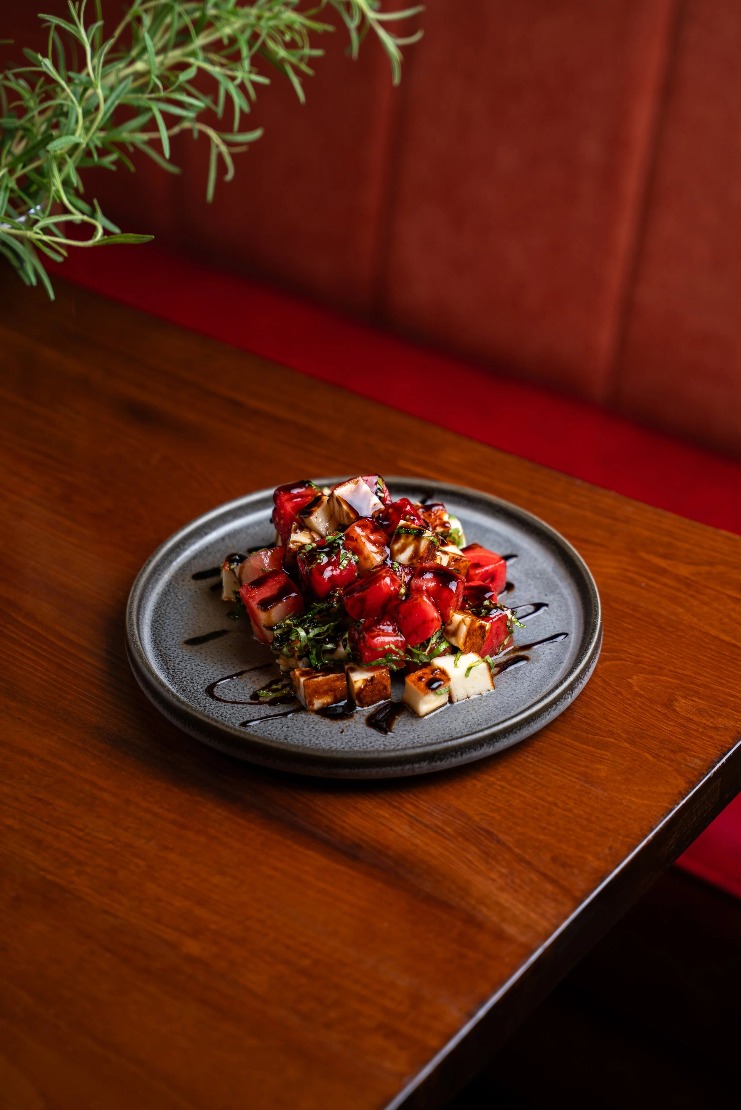 Close-up view of a plate of chopped tofu topped with herbs, drizzle of balsamic reduction, and a glossy sauce, placed on a wooden table with a brown cushioned chair and green plant in the background.