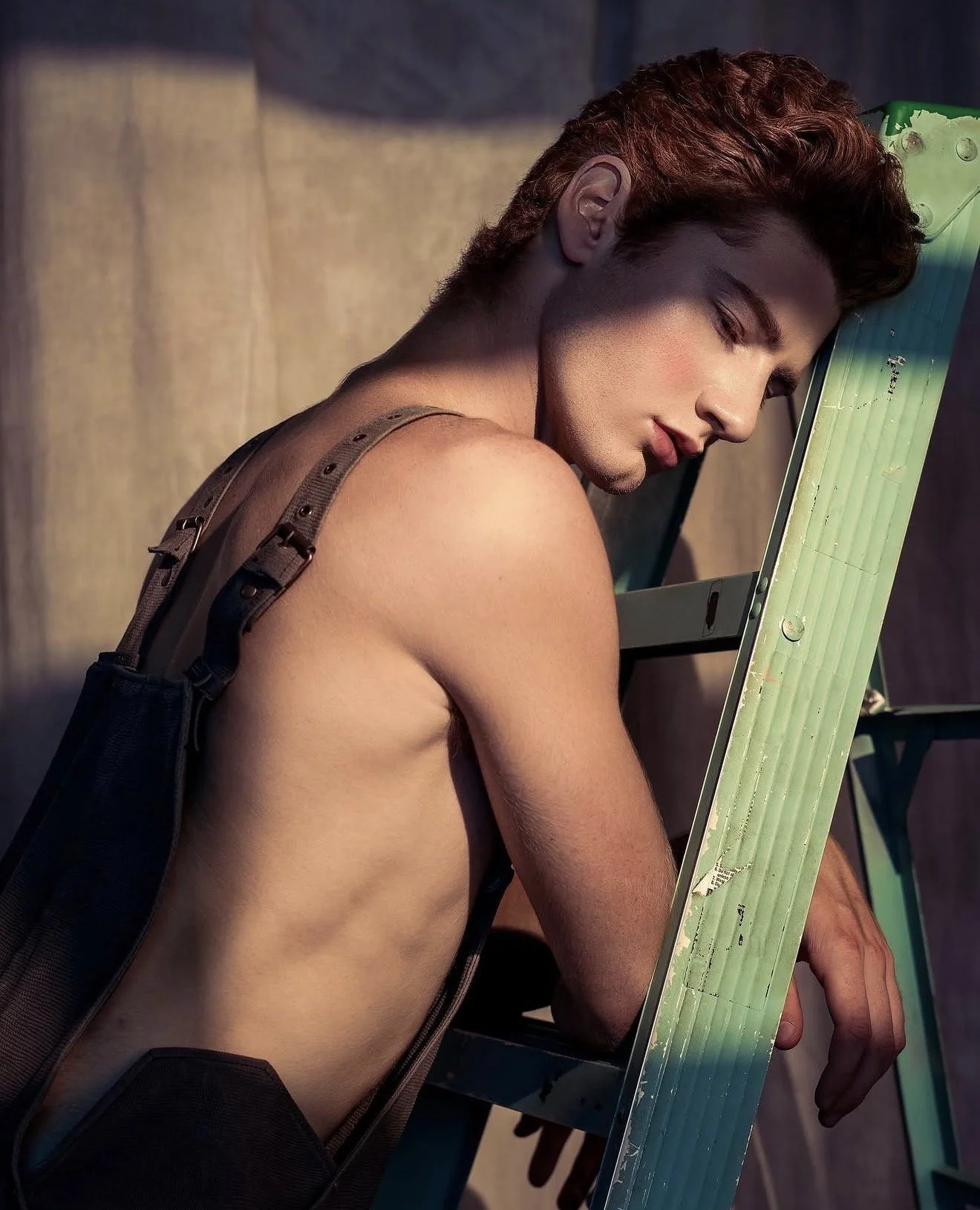 A shirtless young man with curly hair resting his head on a green ladder, with his eyes closed and an expression of peacefulness.
