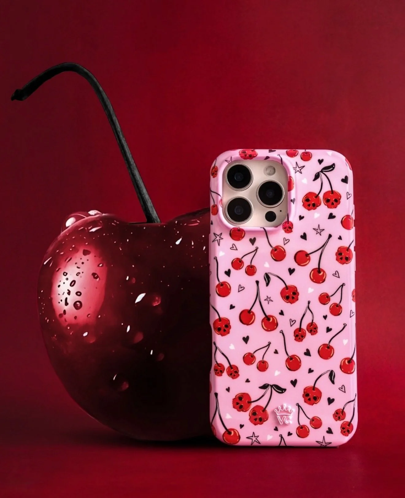 A smartphone with a cherry and skull pattern case placed in front of a large, glossy cherry on a red background.