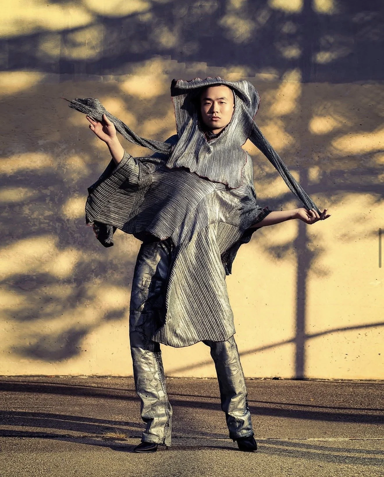 Person dressed in metallic, silver-colored clothing posing outdoors at sunset, with shadows of trees cast on the wall behind him.