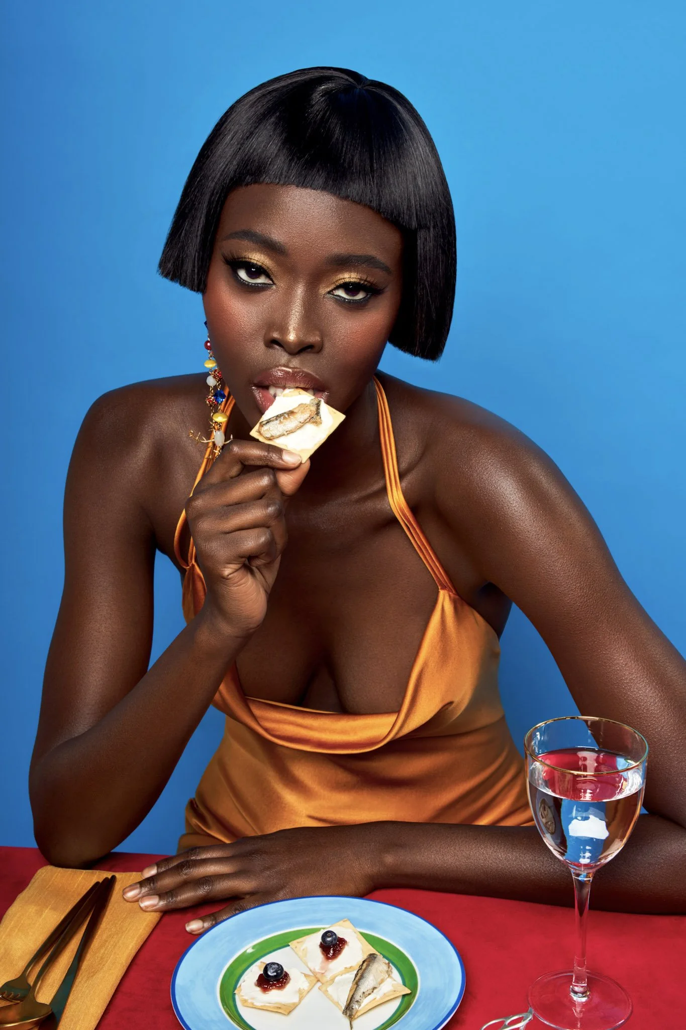 A woman with dark skin and short black hair styled in a blunt bob is sitting at a table. She is wearing gold eyeshadow, earrings with colorful beads, and an orange dress with spaghetti straps. She is holding a piece of cheesecake with blueberry topping near her mouth. In front of her on the table are a plate with four pieces of cheesecake topped with blueberries and jam, a glass of red wine, and a fork and knife on a yellow napkin. The background is blue.