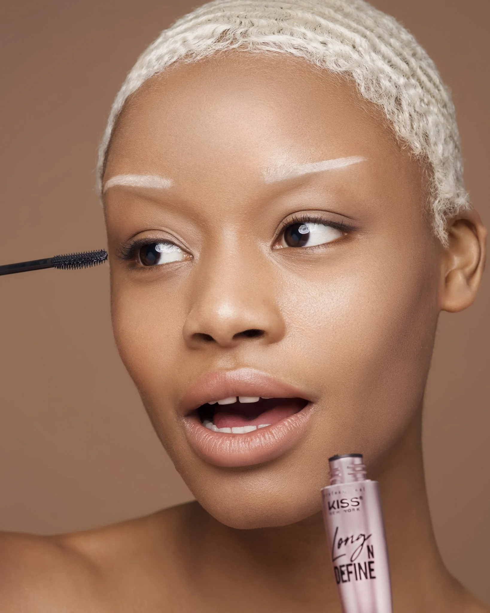 A woman with short platinum blonde hair is applying mascara to her eyelashes while holding a pink lipstick.