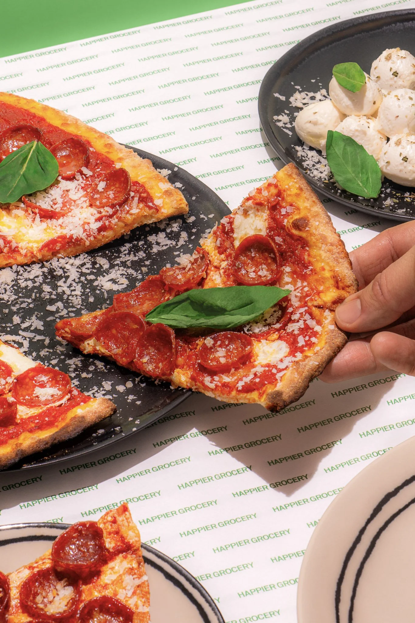 Slices of pepperoni pizza with basil leaves on a black plate and a side of cheese balls garnished with basil leaves on a black plate, on paper with green 'Happier Grocery' printed repeatedly.