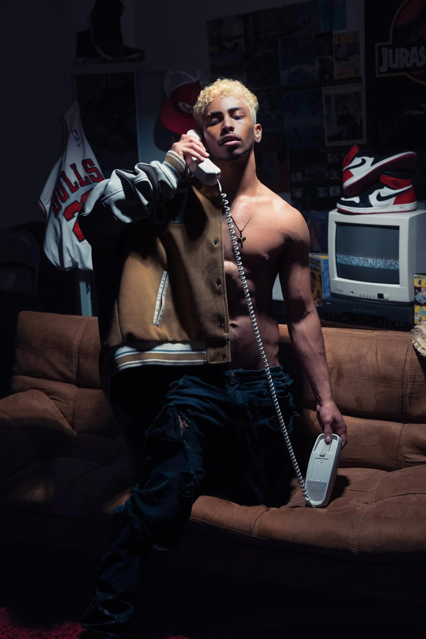 A shirtless young man with dyed blonde hair, wearing a brown varsity jacket and ripped black jeans, stands in a dimly lit room talking on a corded landline phone and holding the receiver in his left hand. There is a vintage TV, a pair of Air Jordan sneakers, a Chicago Bulls jersey, and various posters on the wall behind him.