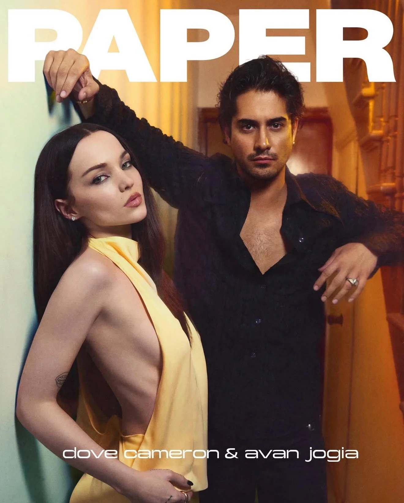 Two models, a woman with long dark hair and a tattoo on her arm wearing a yellow dress and a man with dark hair wearing a black shirt, pose inside a house with warm lighting. The woman leans against a wall, and the man leans against a staircase railing, with his arm resting on it. Large white text at the top says 'PAPER,' and smaller white text at the bottom reads 'dove cameron & avan jogia.'