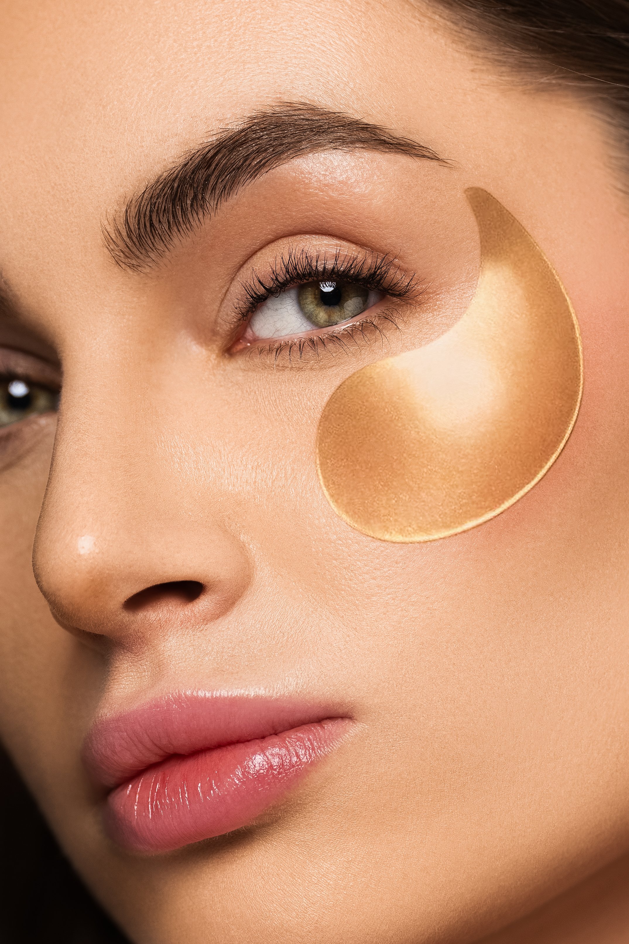 Close-up of a woman's face with a focus on her eye, eyebrow, nose, and lips, highlighting makeup details and skin texture. A gold or bronze patch is under her eye.