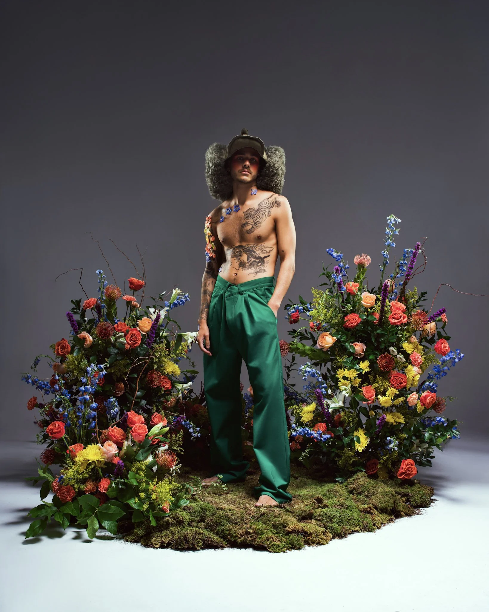 A man with tattoos, wearing green pants and a hat, standing barefoot on a mossy surface surrounded by colorful flowers against a gray backdrop.