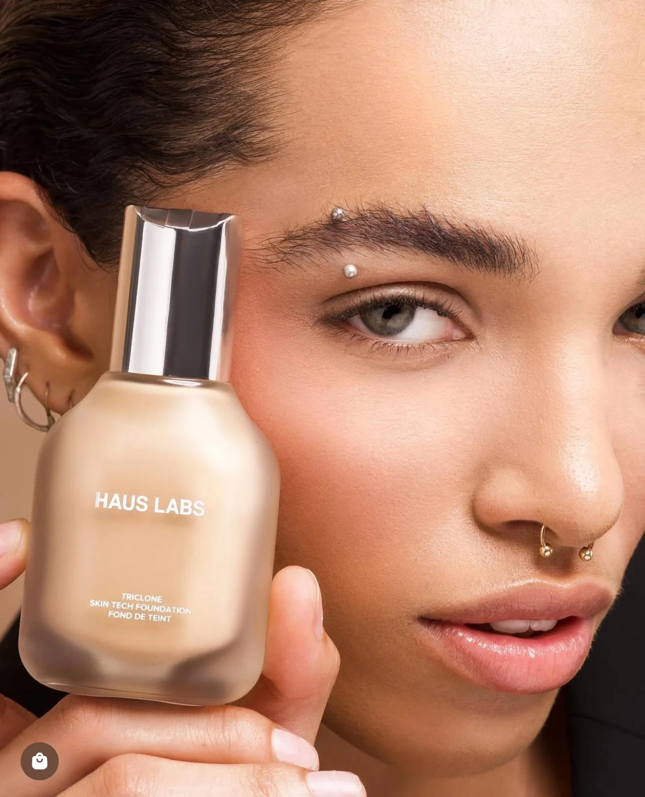 A woman with dark hair and piercings holds a bottle of Haus Labs Triclone Skin Tech Foundation close to her face. The foundation bottle is beige with silver accents.