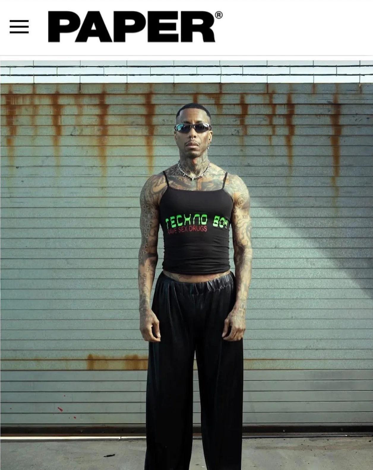 A tattooed man in sunglasses wears a black tank top with the words 'ECHO BOY' and 'DRUG SEX DRUGS' written on it, standing in front of a rusty, metal garage door.