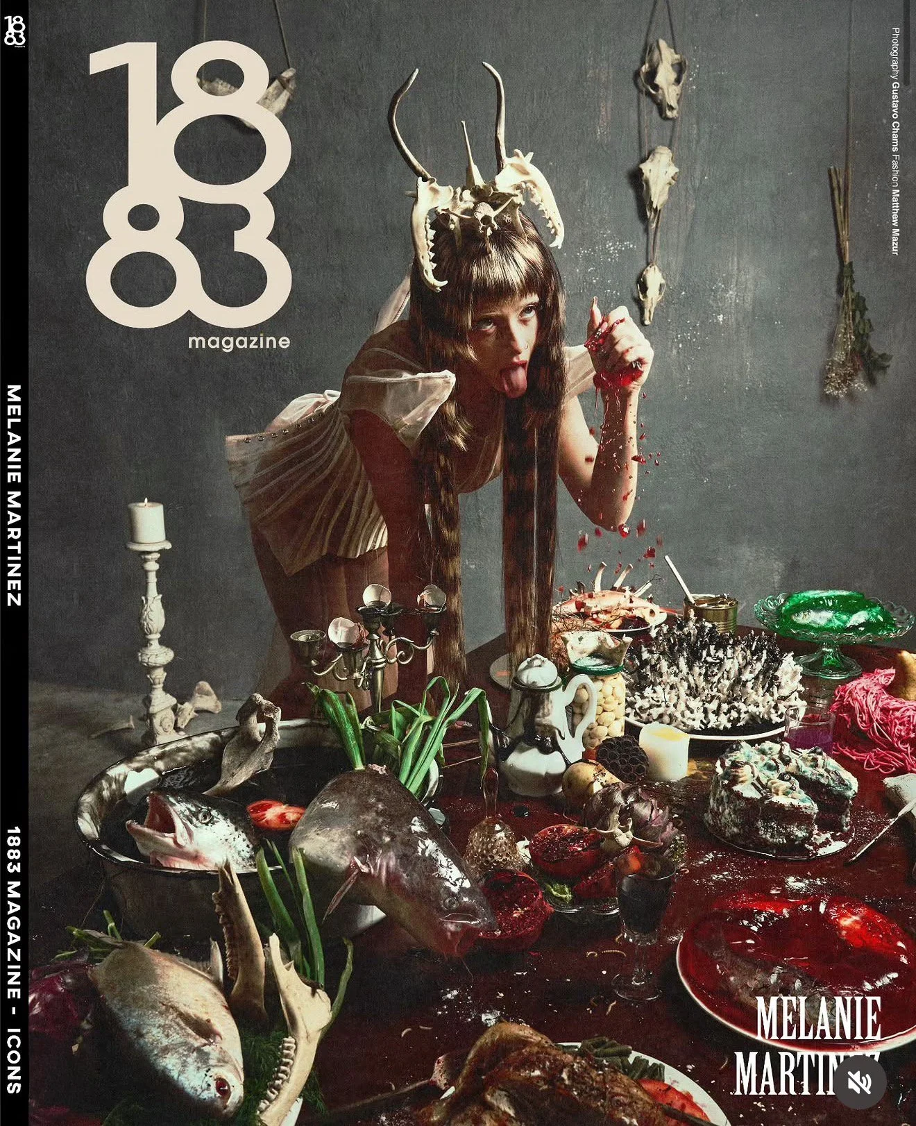 A woman with a skull crown and half-shaved hair leans over a table filled with various food items, including fish, meat, and desserts, in a dark, gothic setting with skull decorations on the wall.